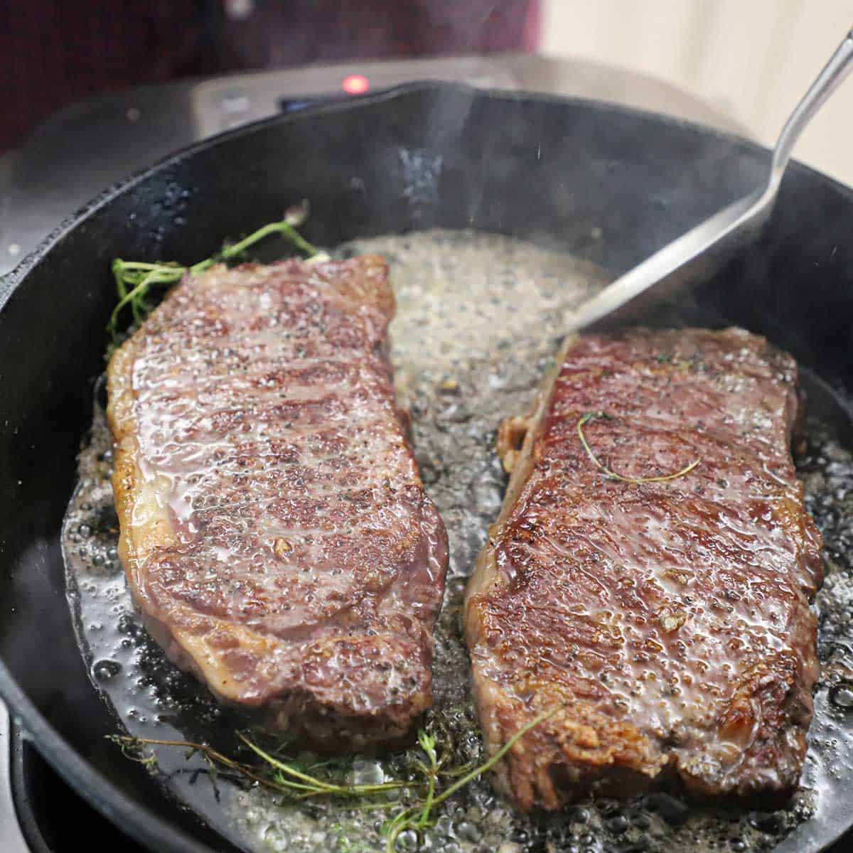 Two New York strip steaks being seared in a very hot cast-iron skillet filled with butter, garlic, and herbs.