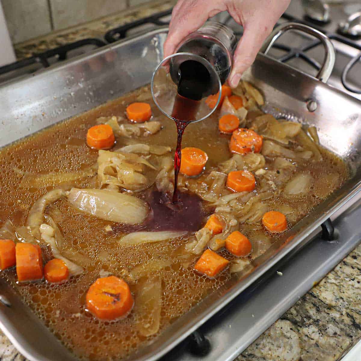 A person pour red wine from a small carafe into a large roasting pan filled with roasted carrots and onions in a beef and lamb stock on a gas stove.