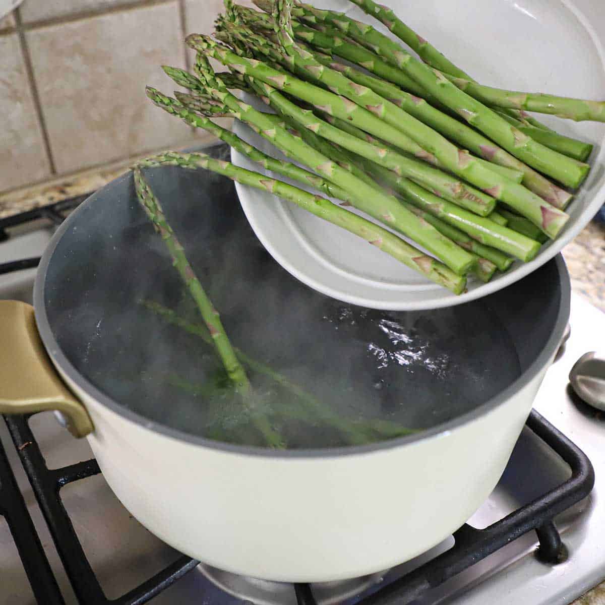 A person transferring a pile of asparagus stalks from a white bowl into a pot filled with boiling water on a gas stove.
