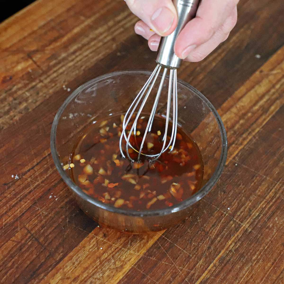 A person using a small whisk to mix a honey garlic glaze in small glass bowl on a wooden cutting board.