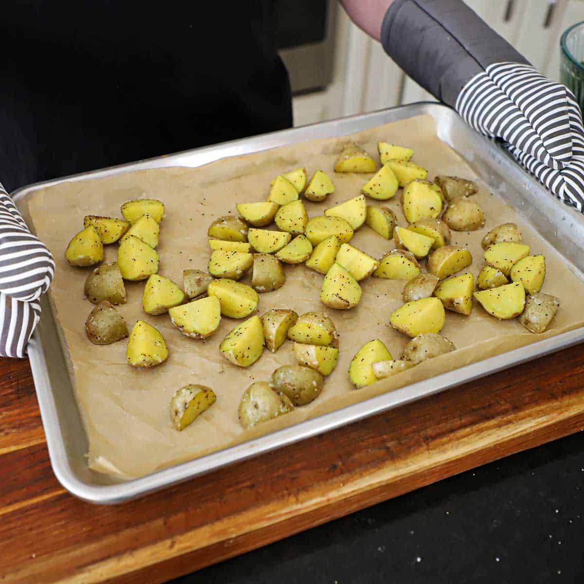 A person using oven mitts to hold a large parchment paper-line sheet pan that is filled with roasted halved baby gold potatoes.