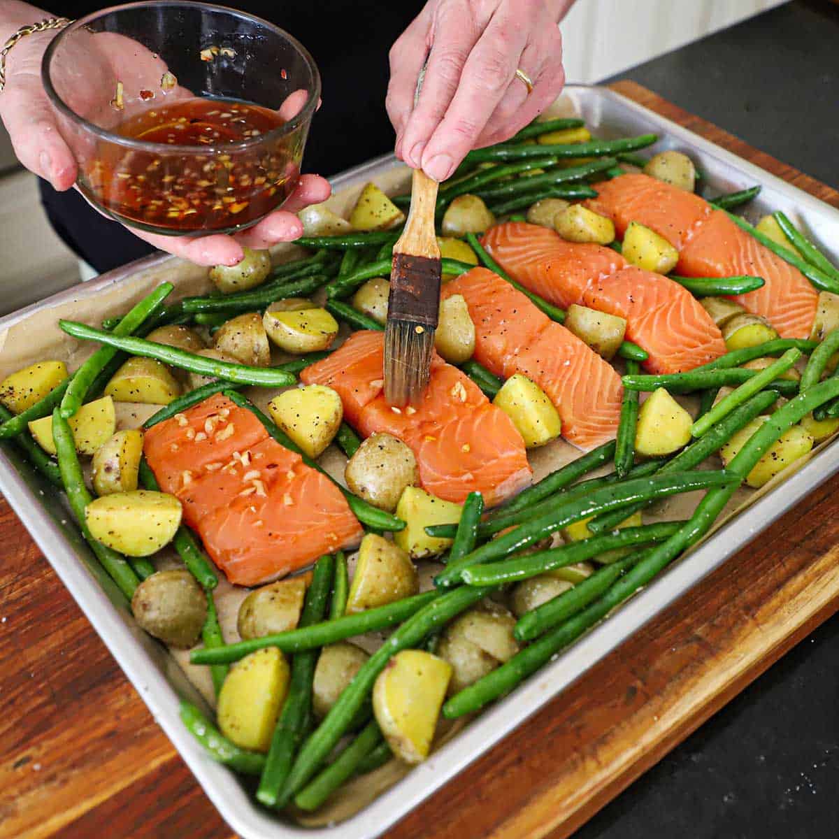 A person using a small brush to apply a honey garlic glaze over the surface of pink salmon filets on a sheet pan nestled with roasted potatoes and uncooked green beans.