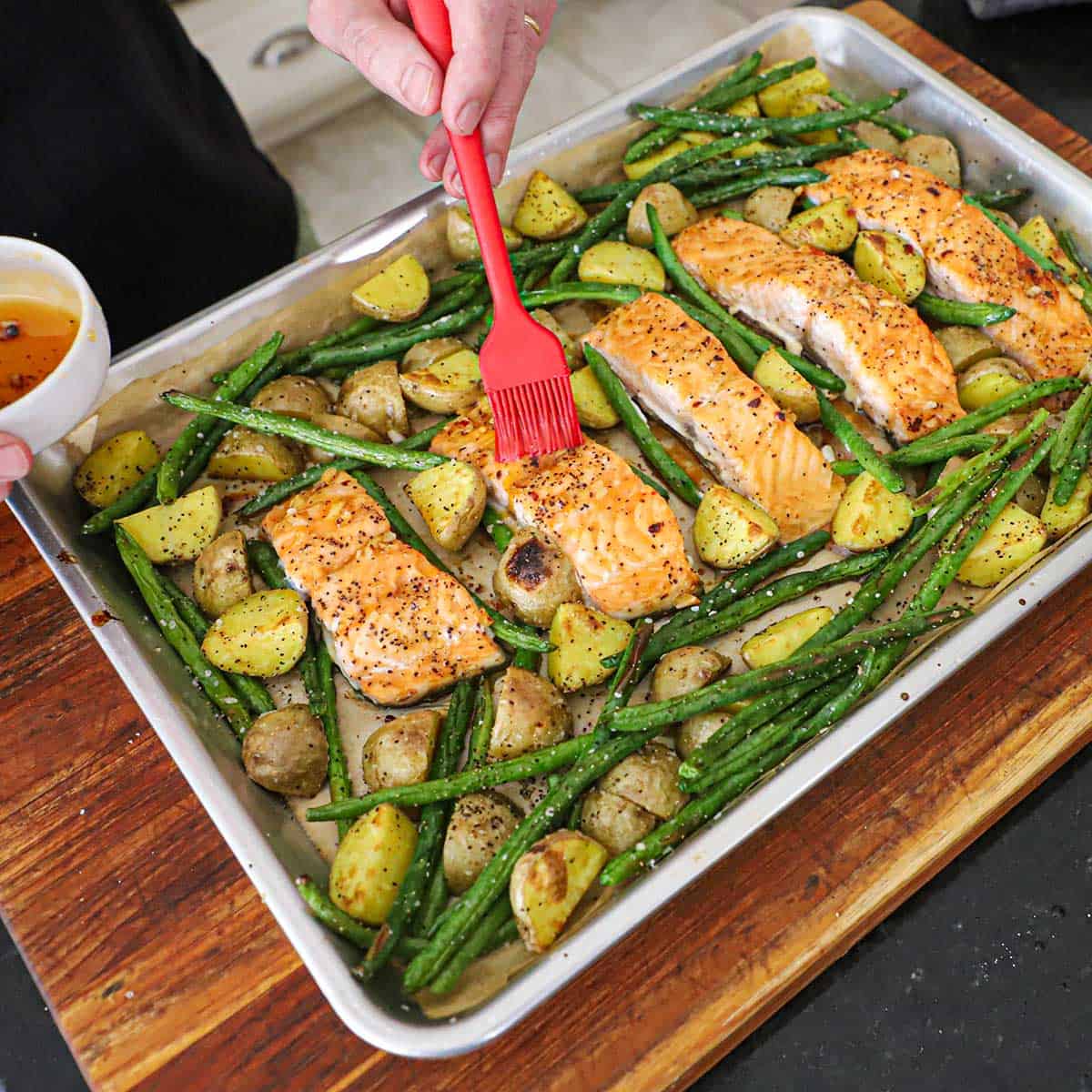 A person using a brush to apply a honey garlic glaze over freshly roasted sheet pan honey garlic salmon and roasted vegetables consisting of green beans and halved baby gold potatoes.