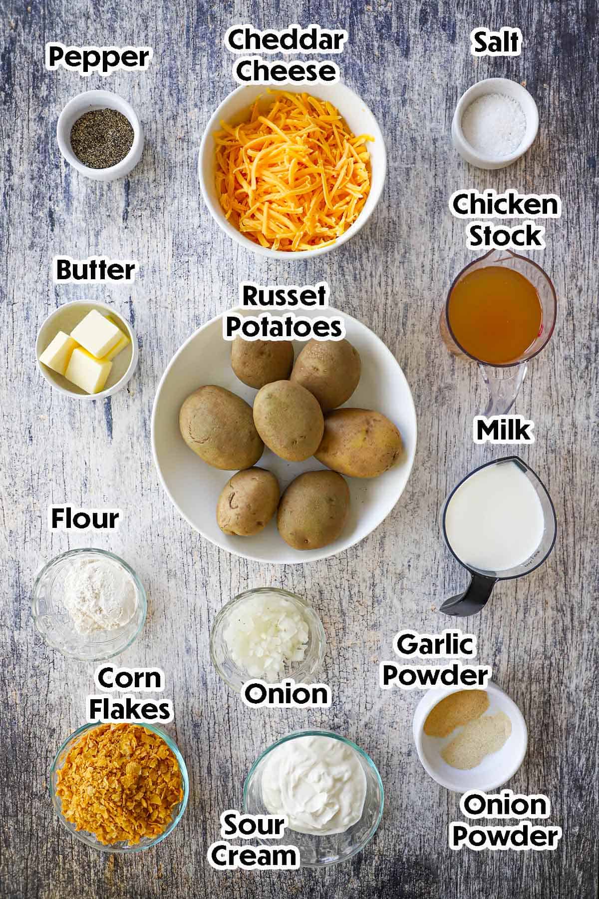 An arrangement of ingredients for hashbrown casserole from scratch on a grey wooden background including russet potatoes, cheddar cheese, chicken stock, butter, milk, flour, onion, corn flakes, sour cream, and seasonings.