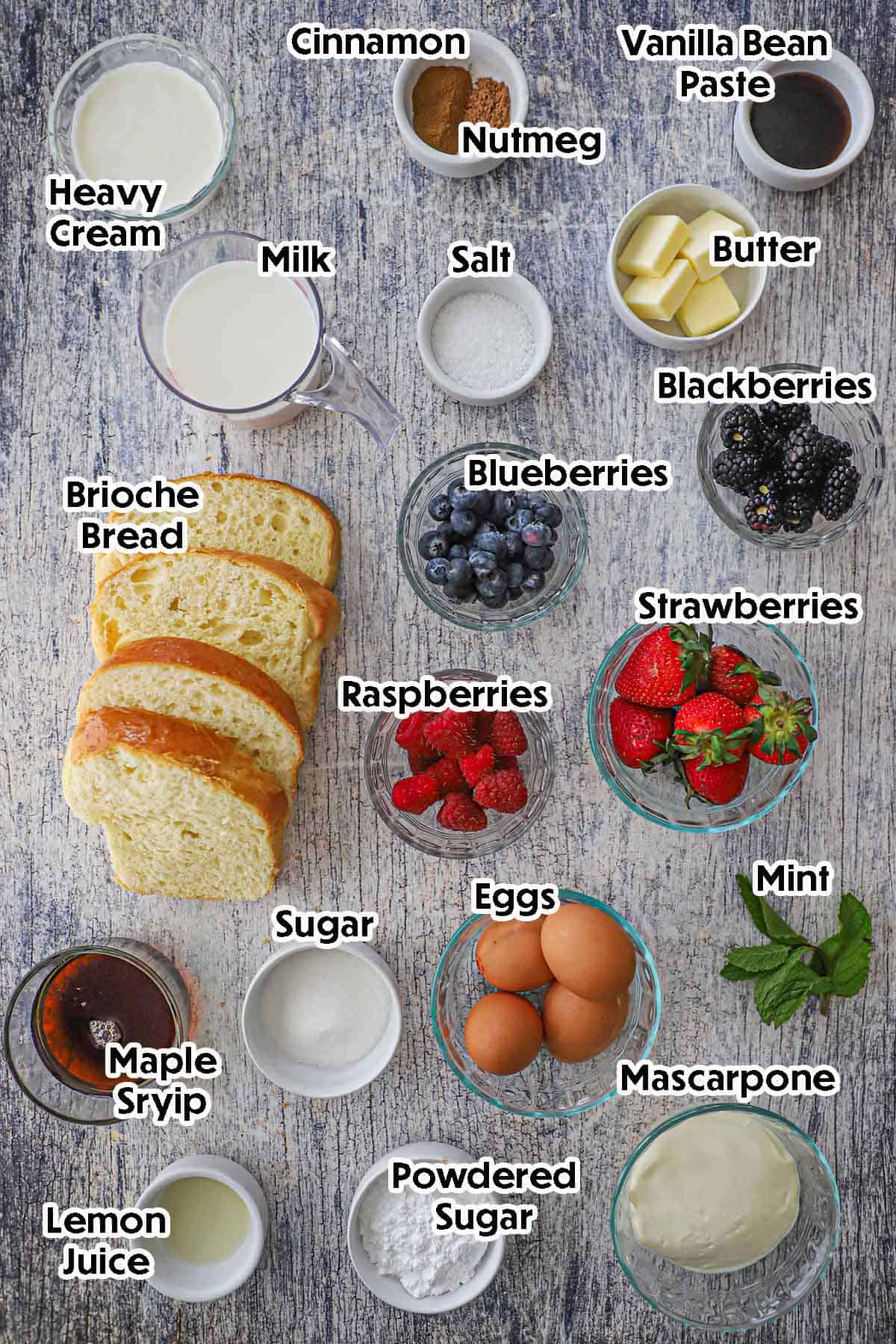 An arrangement of ingredients for Caramelized Brioche French Toast on a grey wooden background including sliced Brioche, eggs, milk, cream, mascarpone, lemon juice, assorted fruits, sugar, and spices.