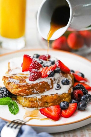 A small white pitcher being used to drizzle warm maple syrup over the top of a plate filled with a serving of caramelized brioche French toast.