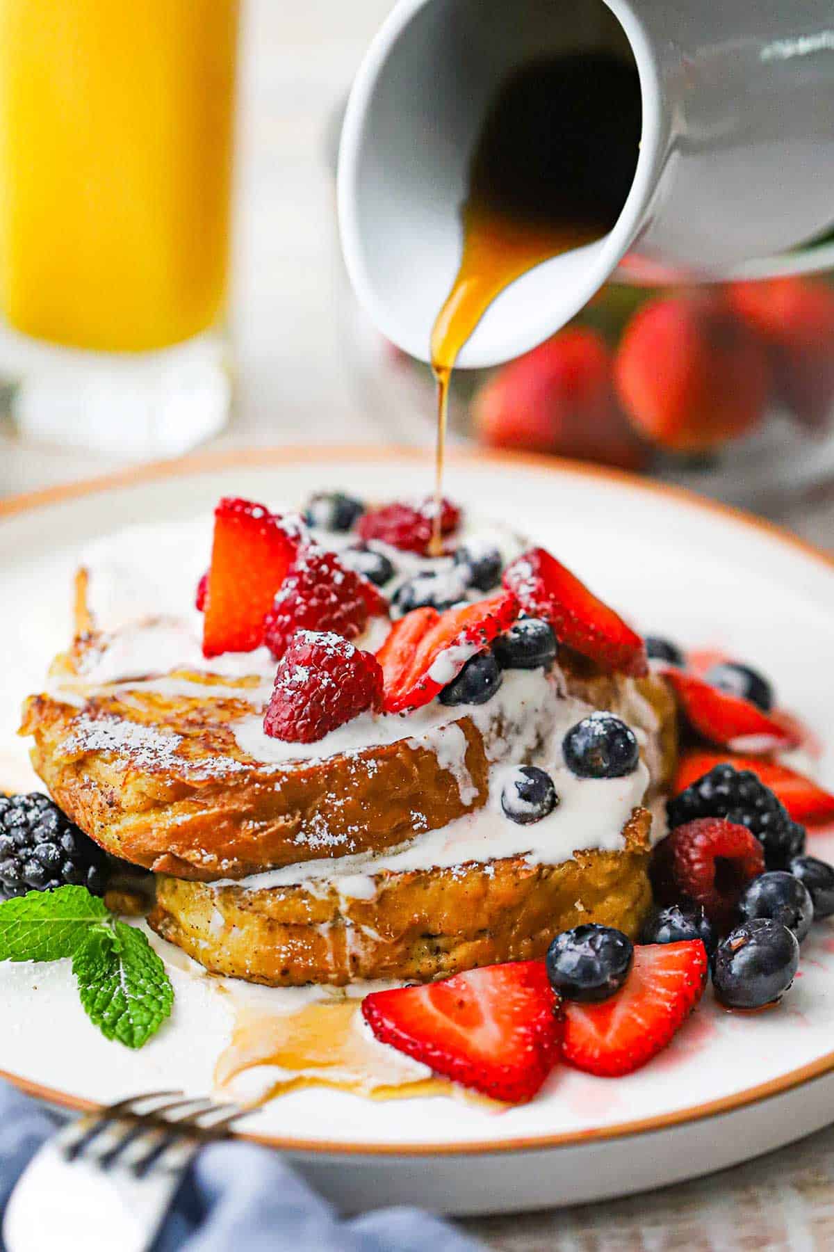 A small white pitcher being used to drizzle warm maple syrup over the top of a plate filled with a serving of caramelized brioche French toast.