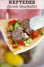 A person using a small spoon to drizzle homemade tzatziki sauce over a split open Greek meatball resting on a pita triangle along with cucumber salad.