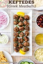 An overhead view of a rectangular white platter filled with two rows of Greek meatballs with small bowls of garnishes nearby.
