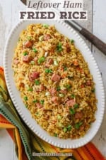 An overhead view of an oval platter containing a large serving of leftover ham fried rice with a colorful napkin and a large serving spoon nearby.