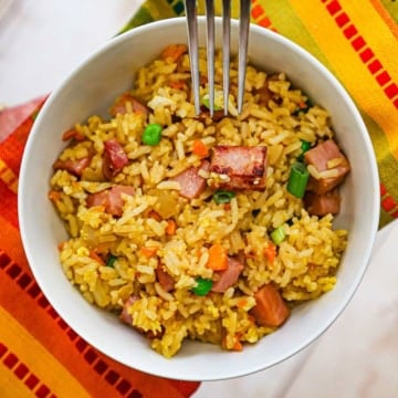 A close-up view of a fork being held over a white bowl filled with a serving of leftover ham fried rice with a colorful napkin underneath the bowl.