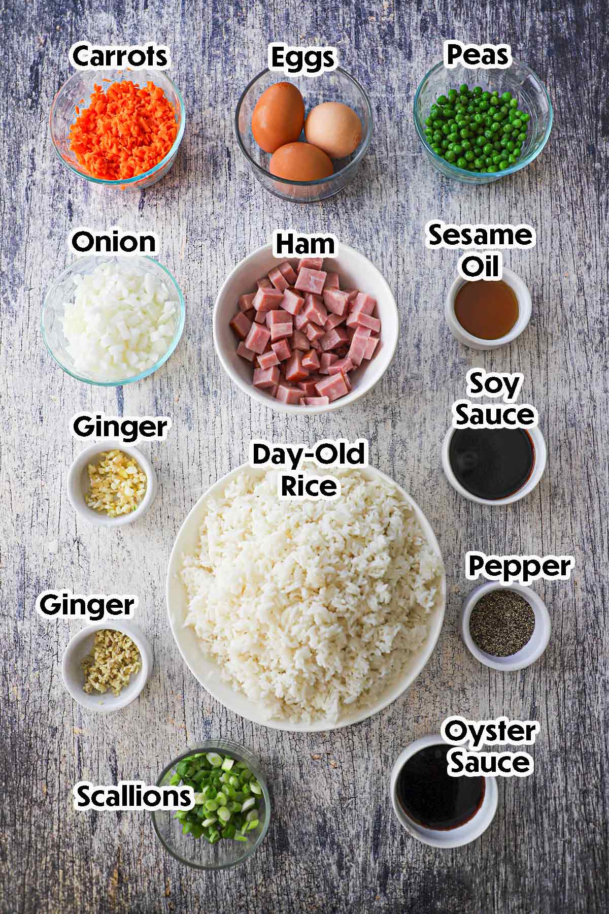 An arrangement of ingredients for leftover ham fried rice on a wooden grey background including cubed ham, day-old rice, carrots, onion, ginger, garlic, peas, scallions, and Chinese seasonings.