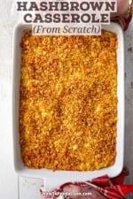 An overhead view of a person using two red-checkered linens to hold the handles of a white baking dish containing an untouched hashbrown casserole from scratch.