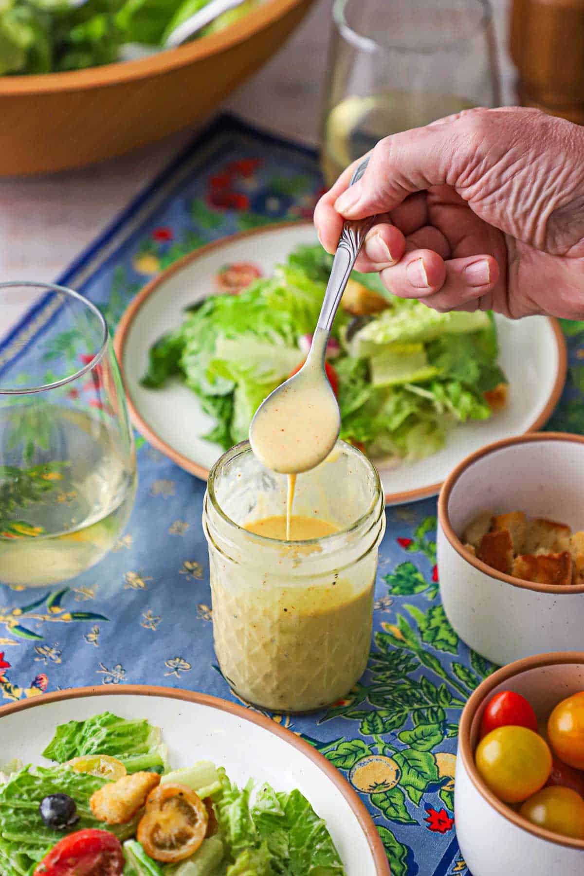 A person holding a spoonful of Restaurant-Style Honey Mustard Dressing over a jar of the same surrounded by a couple of plates holding side salads as well as a glass of white wine a small jar holding cherry tomatoes.