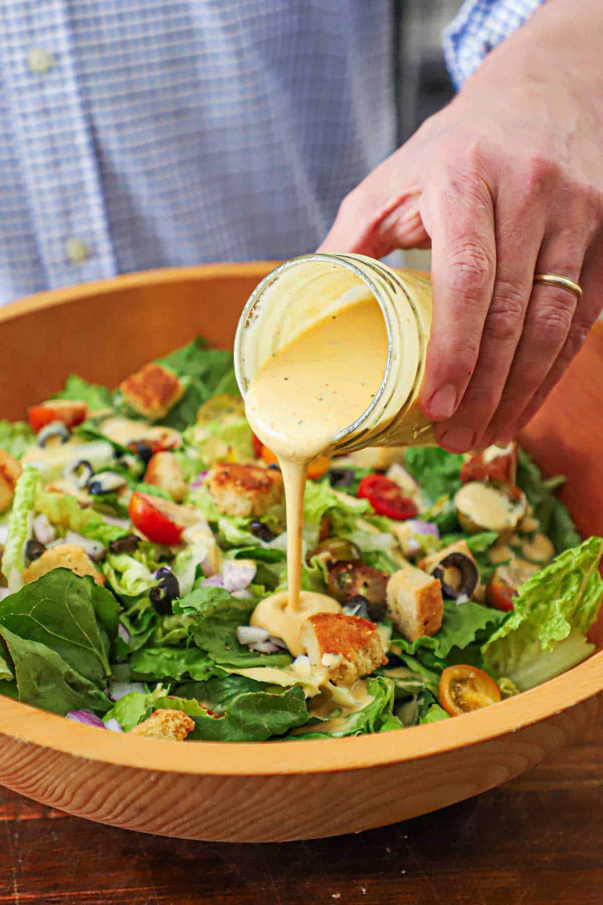 A person pouring Restaurant-Style Honey Mustard Dressing from a jar over a chopped salad in a large wooden salad bowl
