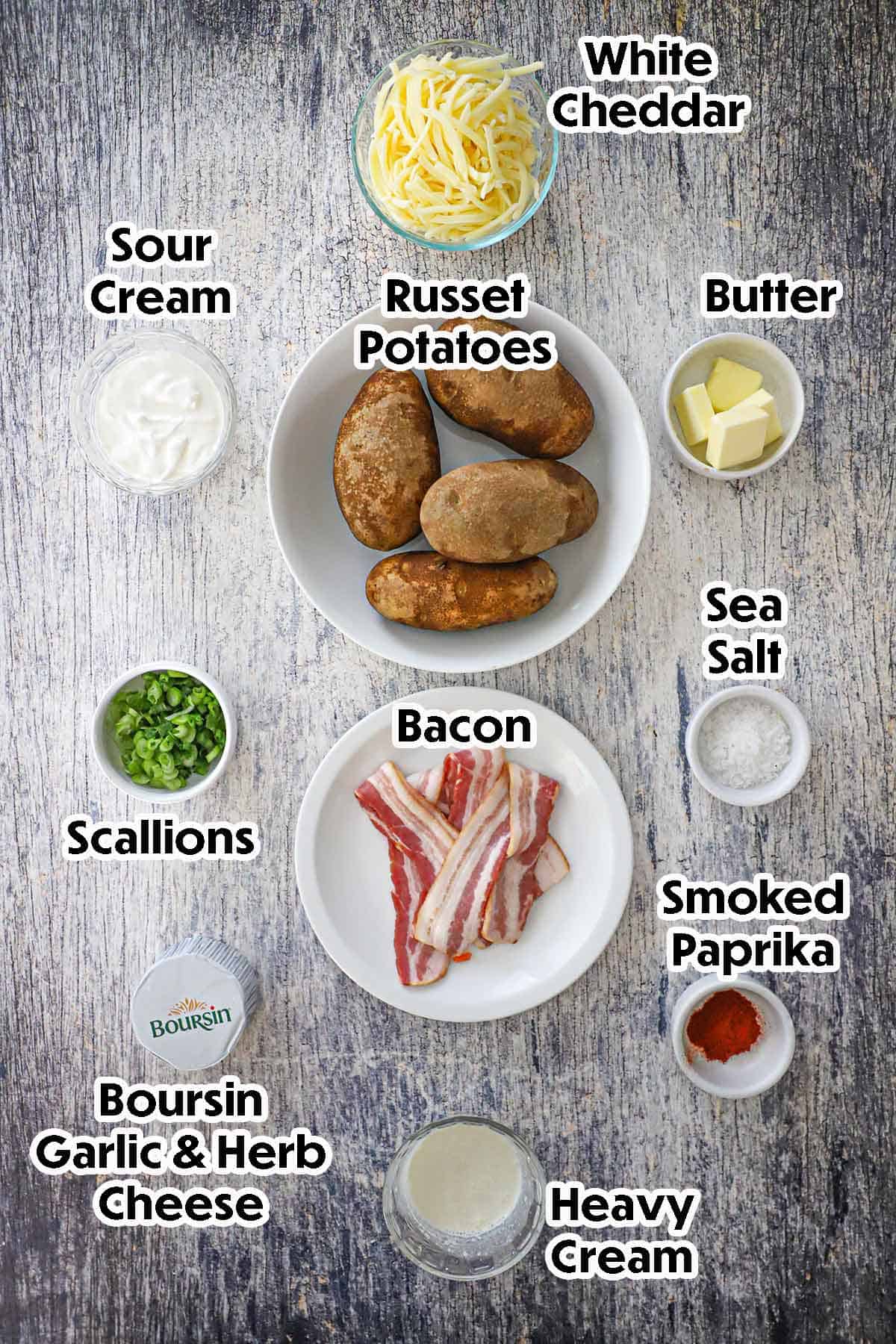 An arrangement of ingredients for twice baked potatoes on a grey wooden background including russet potatoes, sour cream, garlic and herb cheese, cream, white cheddar, butter, and seasonings.