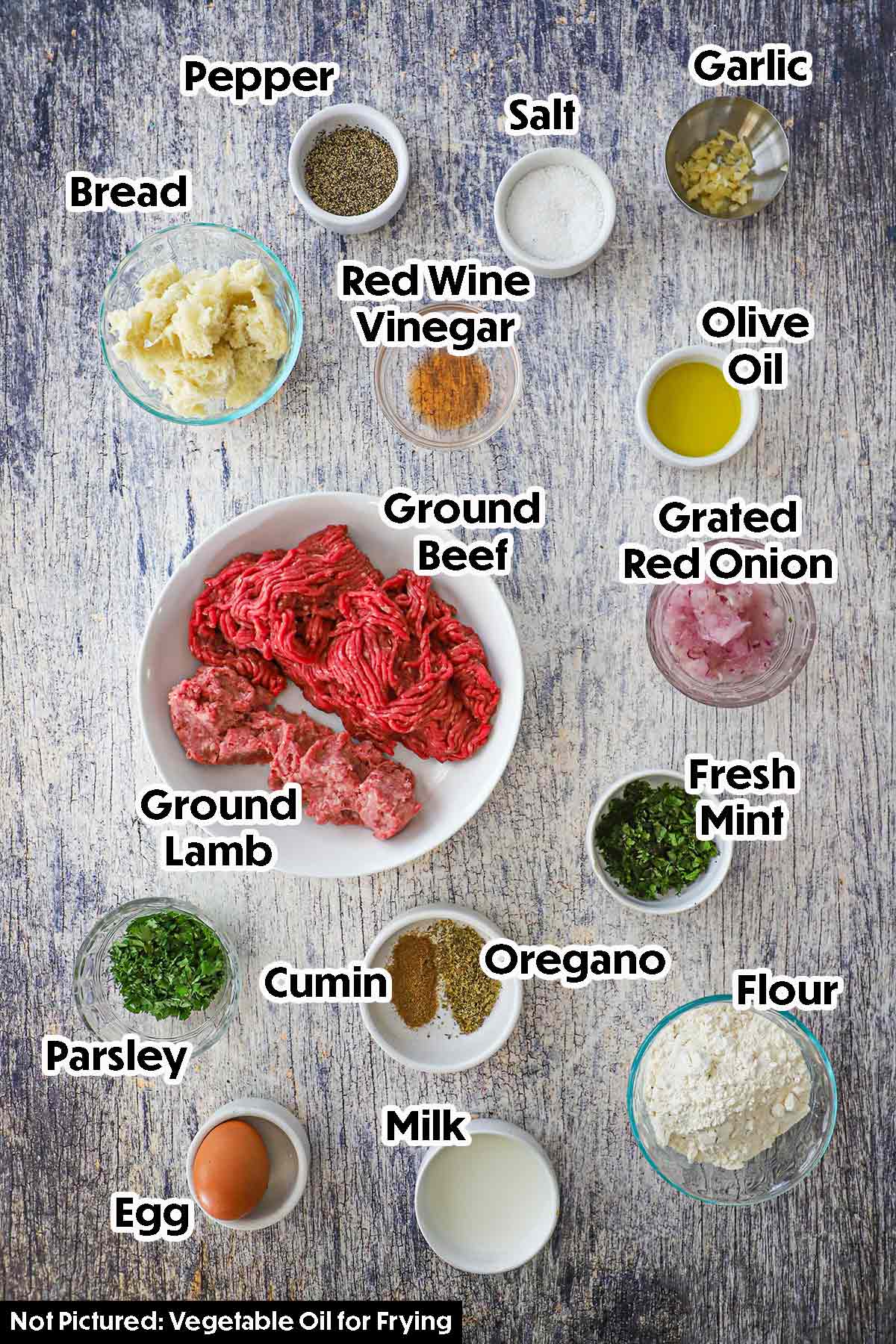 An arrangement of ingredients for Greek meatballs on a grey wooden background including ground beef, ground lamb, bread, milk, garlic, vinegar, grated onion, chopped herbs, egg, flour, and seasonings.