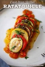 A slanted row of thinly sliced vegetables from a cast-iron skillet ratatouille resting on a thin bed of sautéed tomatoes and red bell pepper all on a white dinner plate.