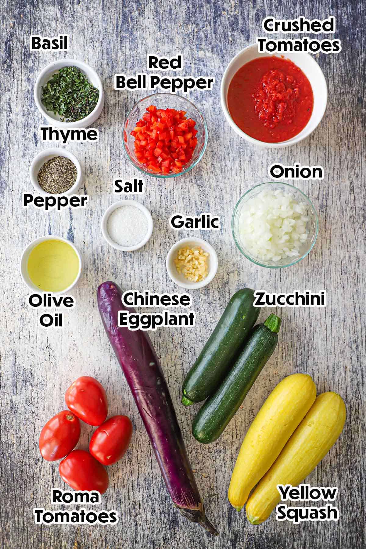An arrangement of ingredients for cast-iron skillet ratatouille on a grey wooden background including zucchini, yellow squash, tomatoes, chopped onion, Chinese eggplant, red bell pepper, crush tomatoes, herbs, and seasonings.