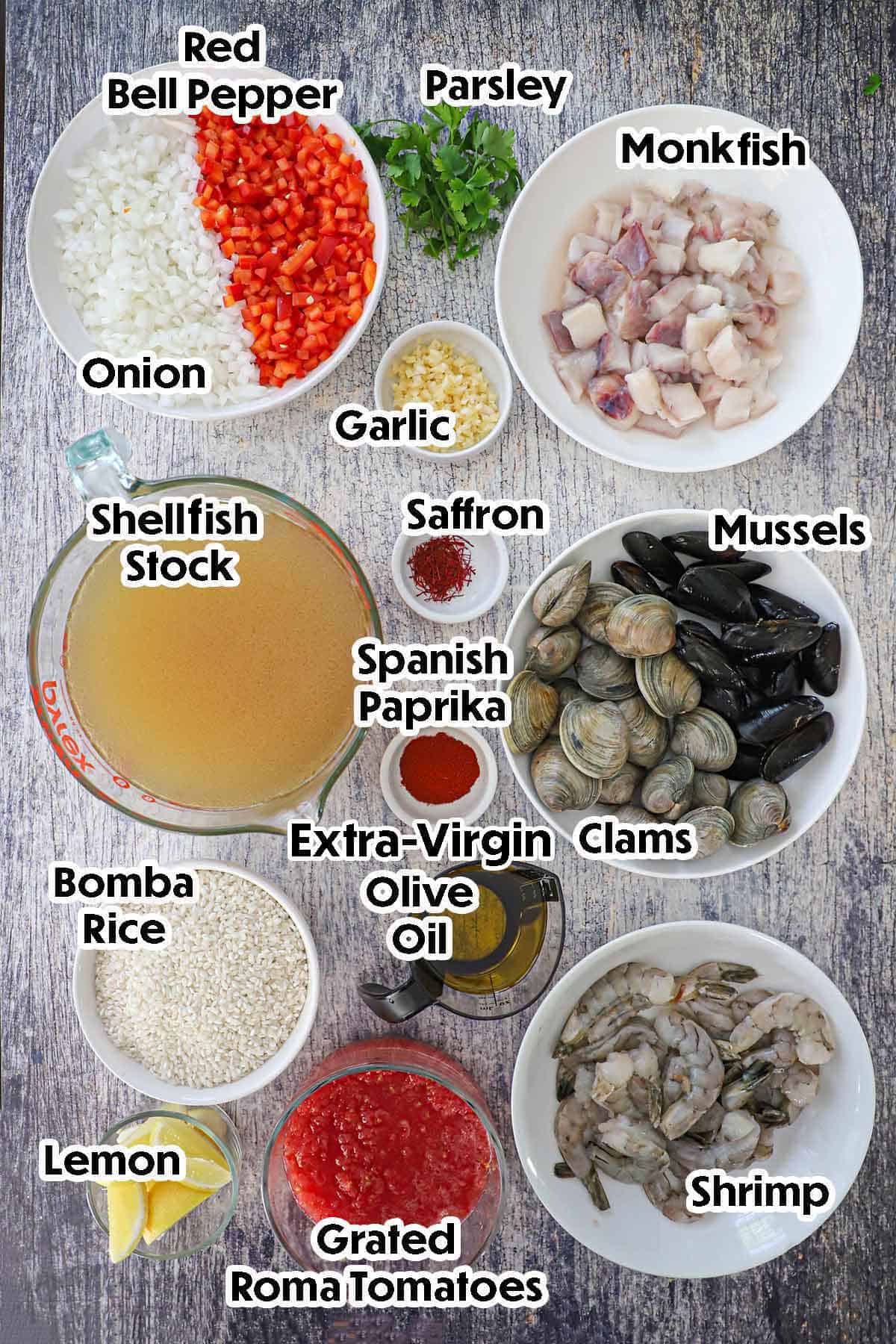 An arrangement of ingredients for Authentic Spanish Seafood Paella on a grey wooden background including bomba rice, fish stock, chopped bell peppers and onions, monk fish, mussels, clams, shrimp, grated tomatoes, garlic, and seasonings.