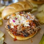 A close-up view of a smoked pulled pork sandwich resting on brown sandwich paper with shredded pork, BBQ sauce, and coleslaw all nestled between two toasted hamburger buns.
