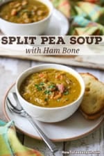 Two white soup bowls filled with split pea soup with ham bone on small plates also holding a slice of toasted bread and a soup spoon.