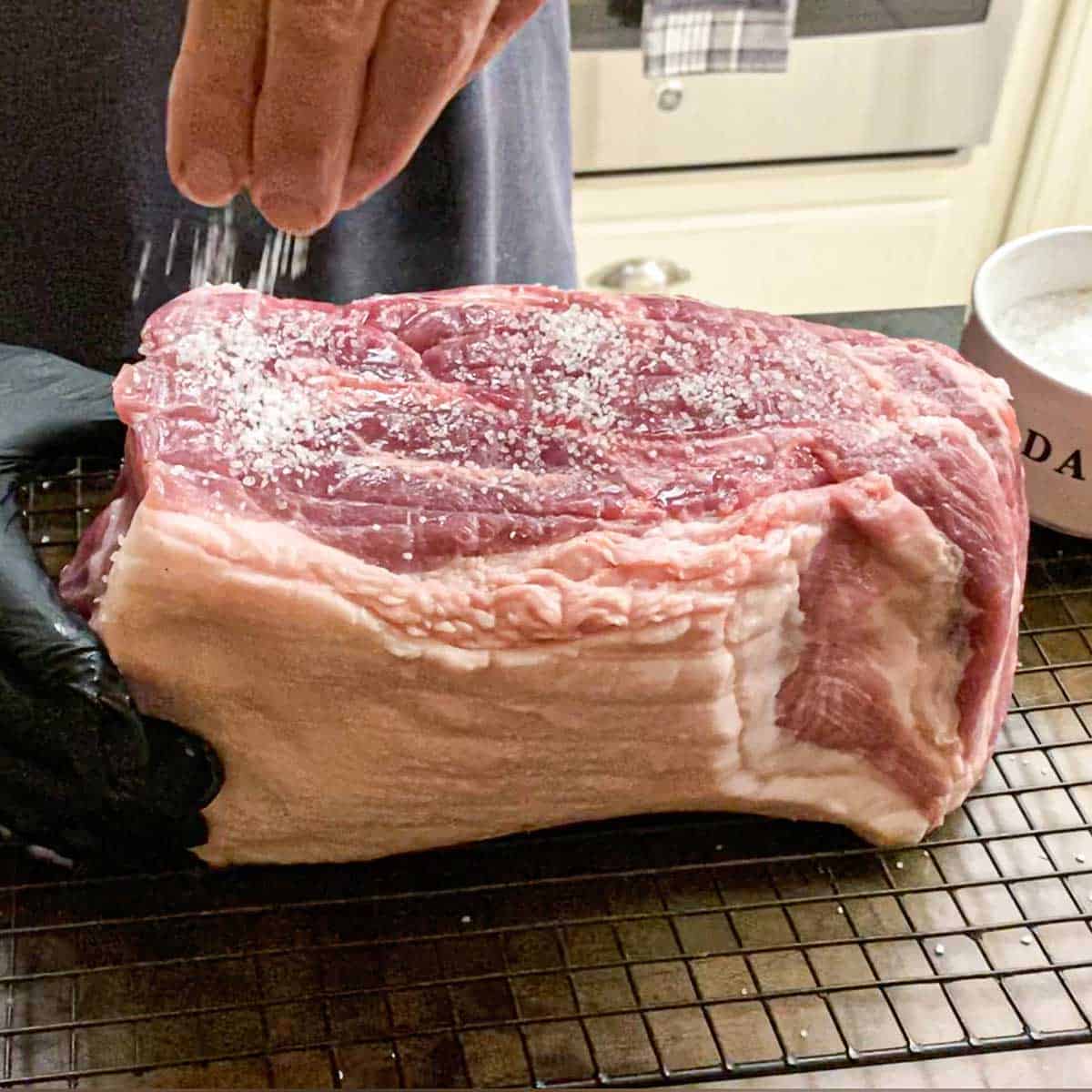 A person sprinkling coarse Koser salt all over an uncooked pork butt that is resting on a baking rack.