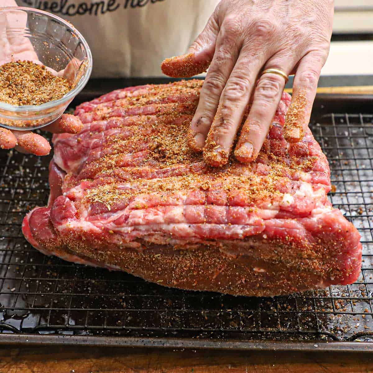 A person using his fingers to press a pork rub into the surface of an uncooked pork butt that is on a baking rack inside a baking sheet pan.