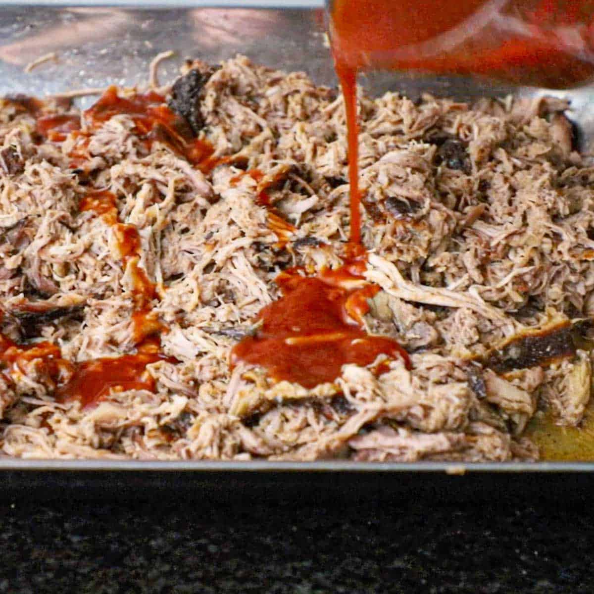 A person drizzling Carolina BBQ sauce over shredded smoked pork in a large steel roasting pan.