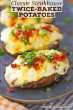 A row of freshly prepared twice baked potatoes topped with chopped scallions and crispy bacon bits all on a grayish-blue oval platter.
