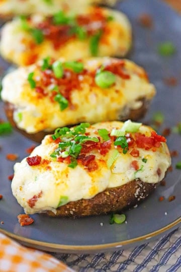 A row of freshly prepared twice baked potatoes topped with chopped scallions and crispy bacon bits all on a grayish-blue oval platter.