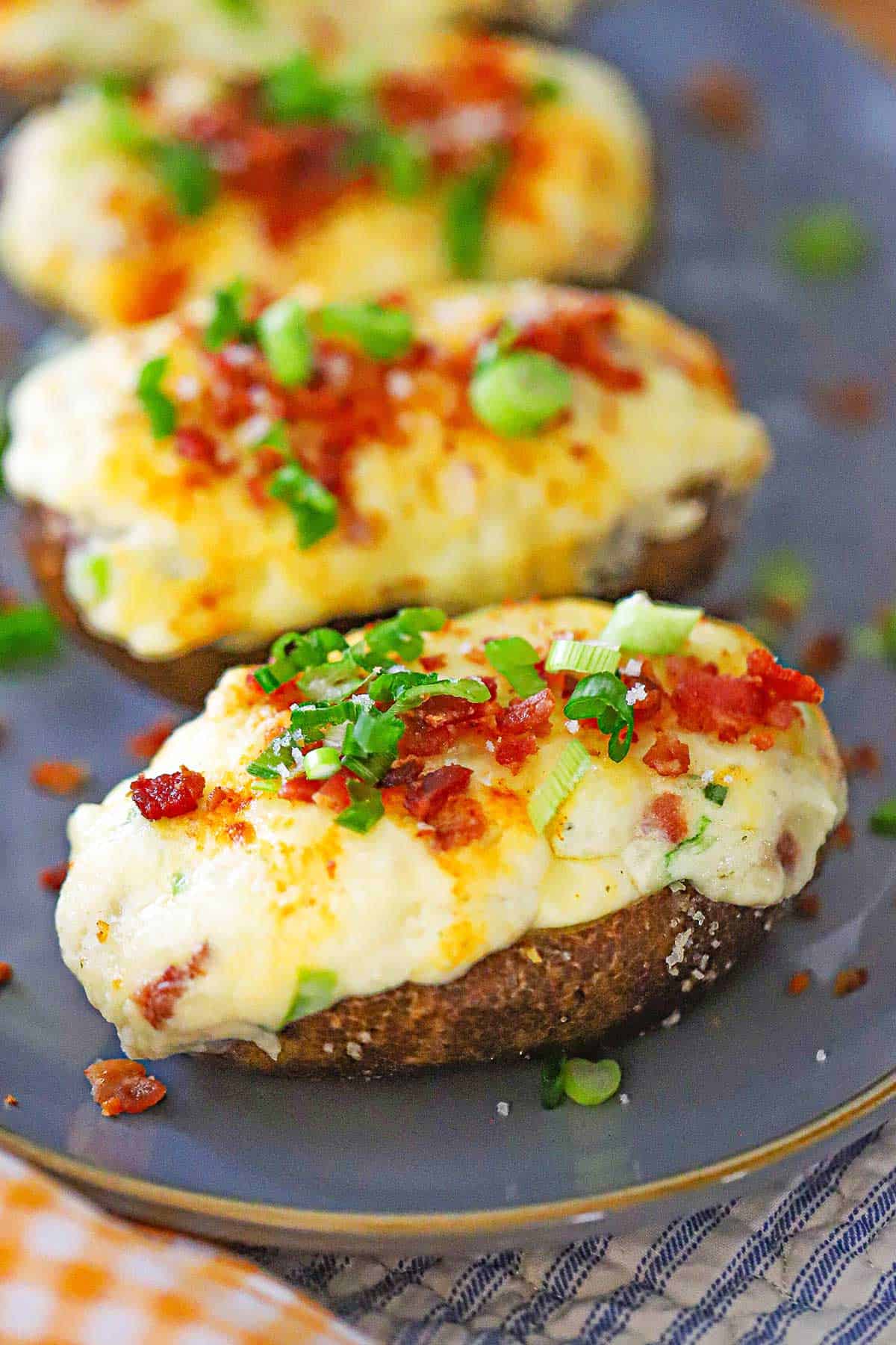 A row of freshly prepared twice baked potatoes topped with chopped scallions and crispy bacon bits all on a grayish-blue oval platter.