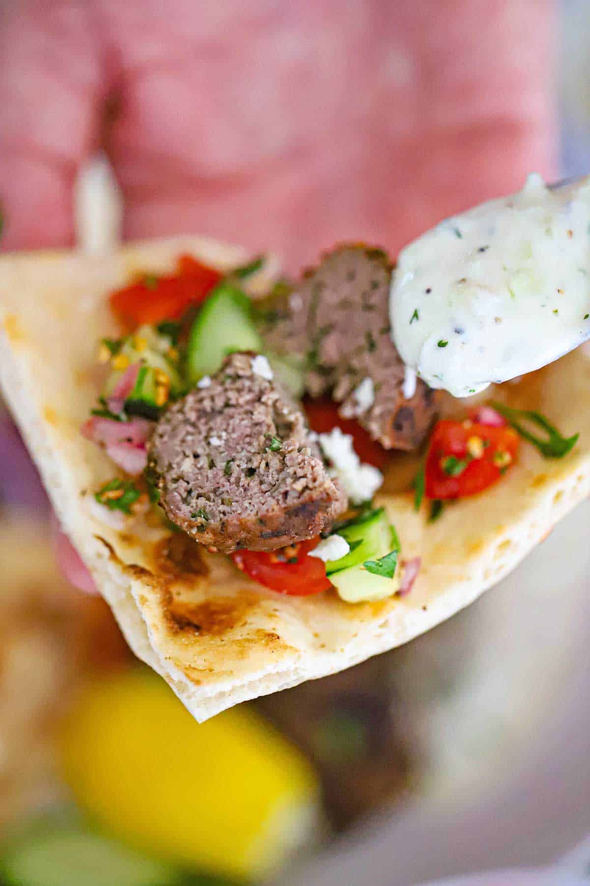 A person using a small spoon to drizzle homemade tzatziki sauce over a split open Greek meatball resting on a pita triangle along with cucumber salad.