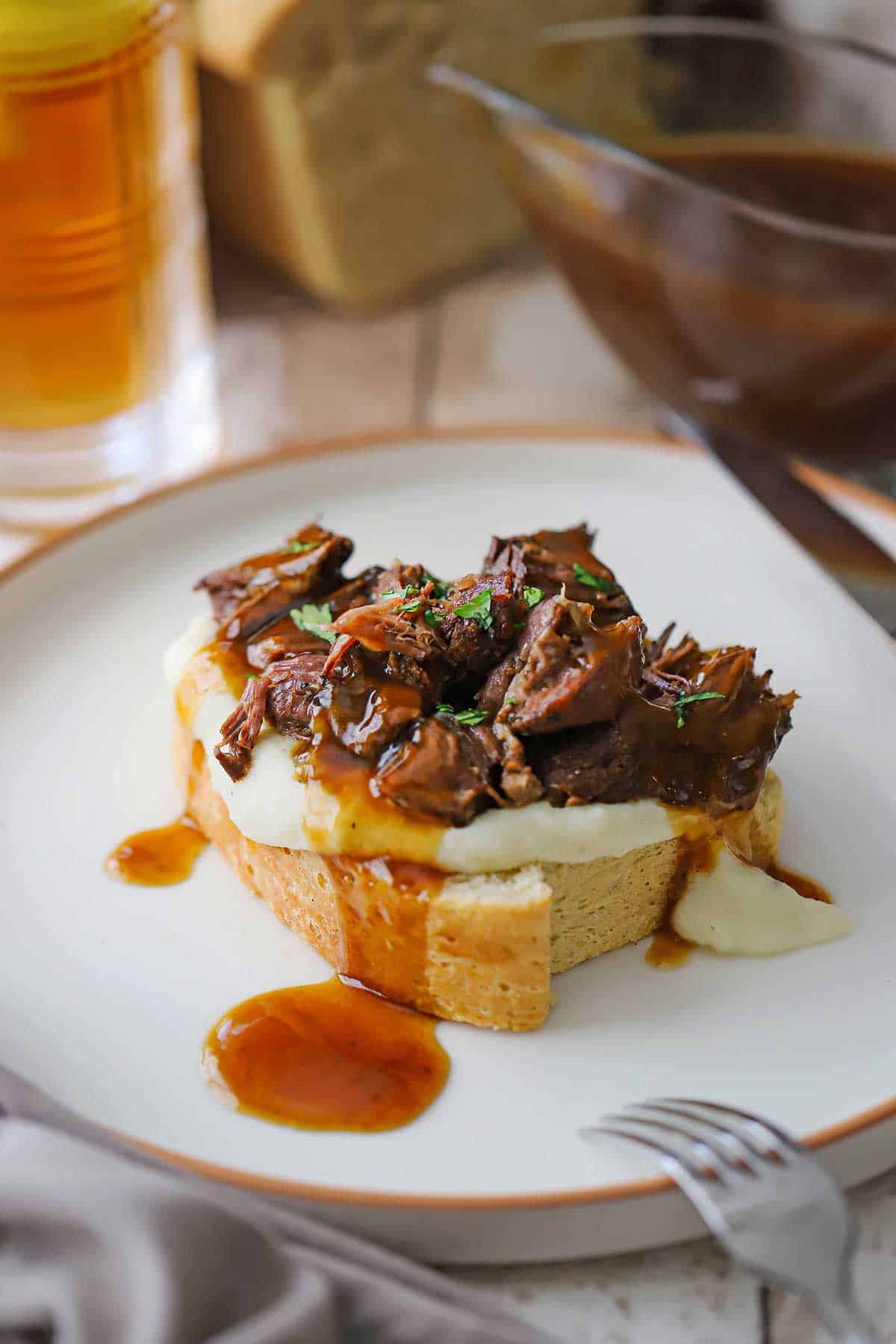 A straight-on view of a slow cooker beef manhattan with brown gravy drizzled over the top of the bread, potatoes, and shredded roasted beef all on a white dinner plate.