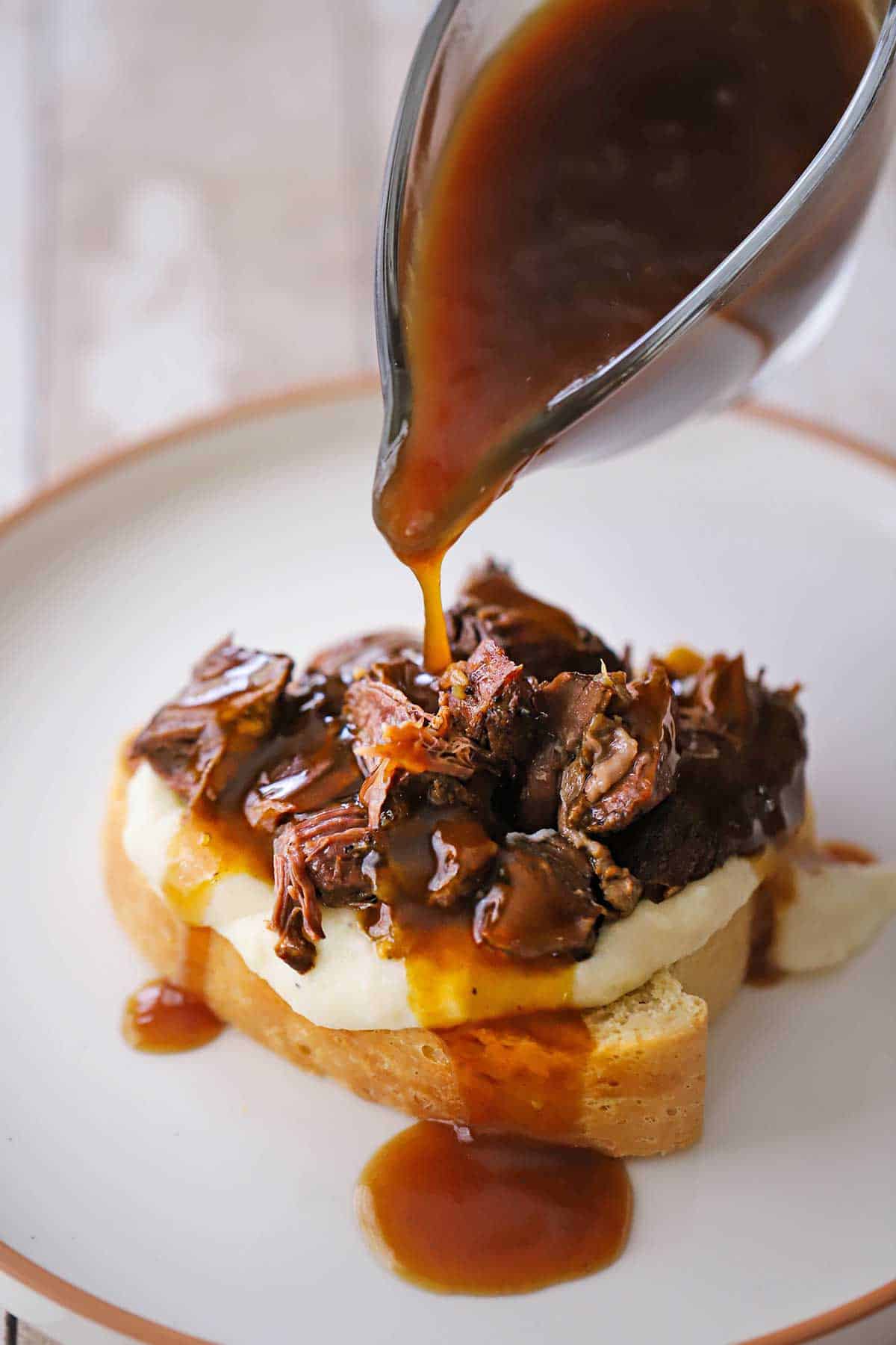 A person pouring brown gravy from a glass gravy boat over the top of a serving of slow cooker beef Manhattan on a white dinner plate.