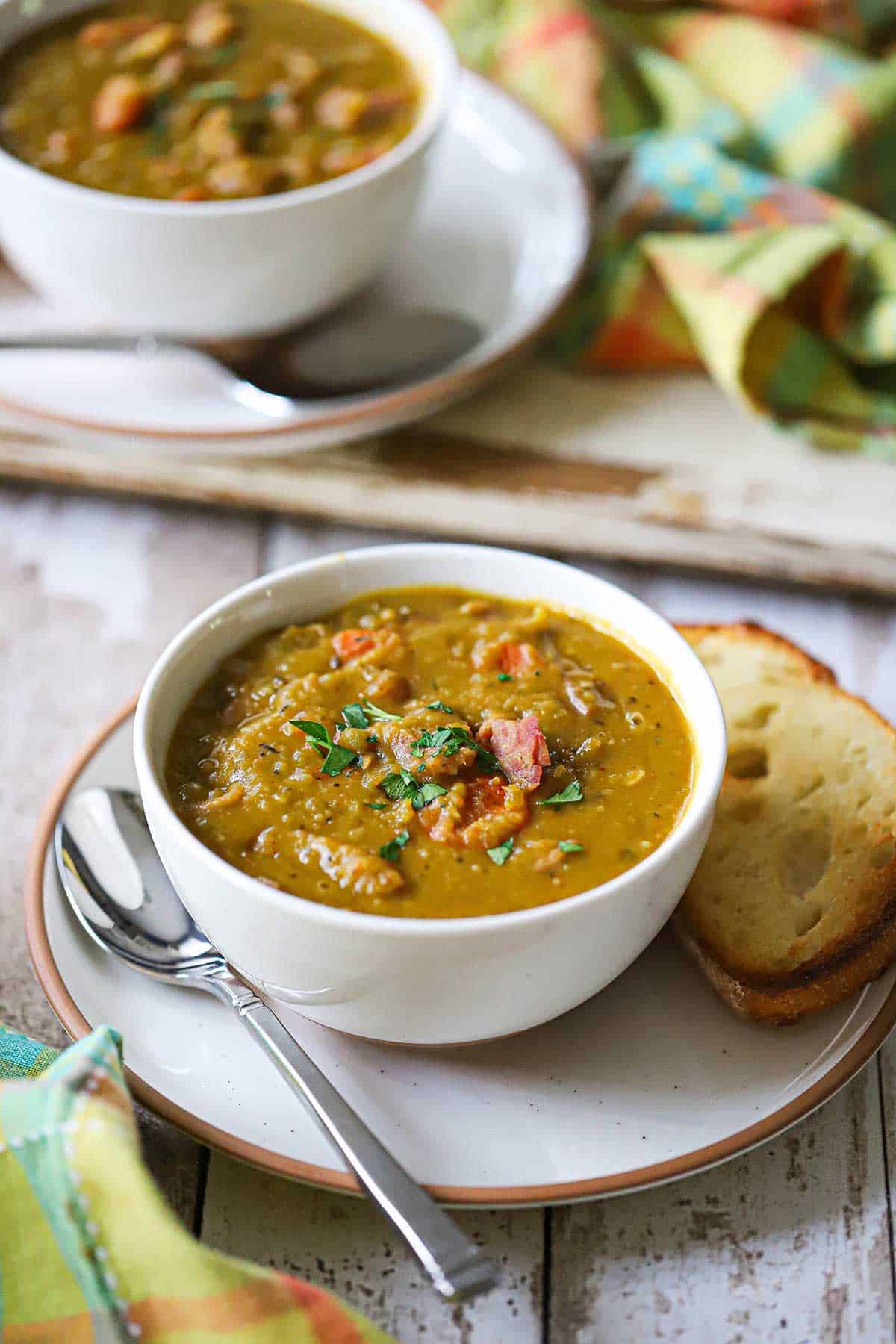 Two white soup bowls filled with split pea soup with ham bone on small plates also holding a slice of toasted bread and a soup spoon.