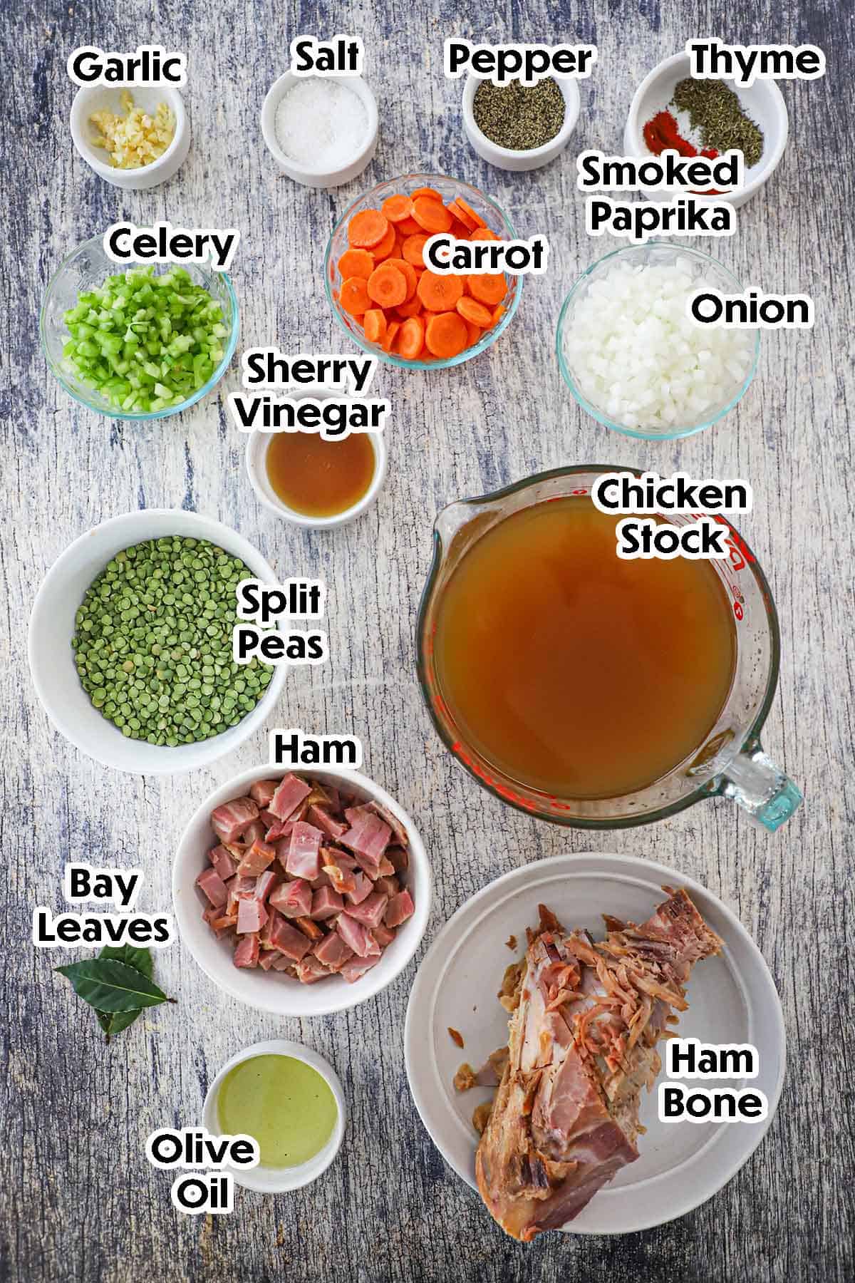 An arrangement of ingredients for split pea soup with ham bone on grey wooden background including split peas, celery, carrots, onion, chicken stock, cubed ham, a ham bone, garlic, and seasonings.