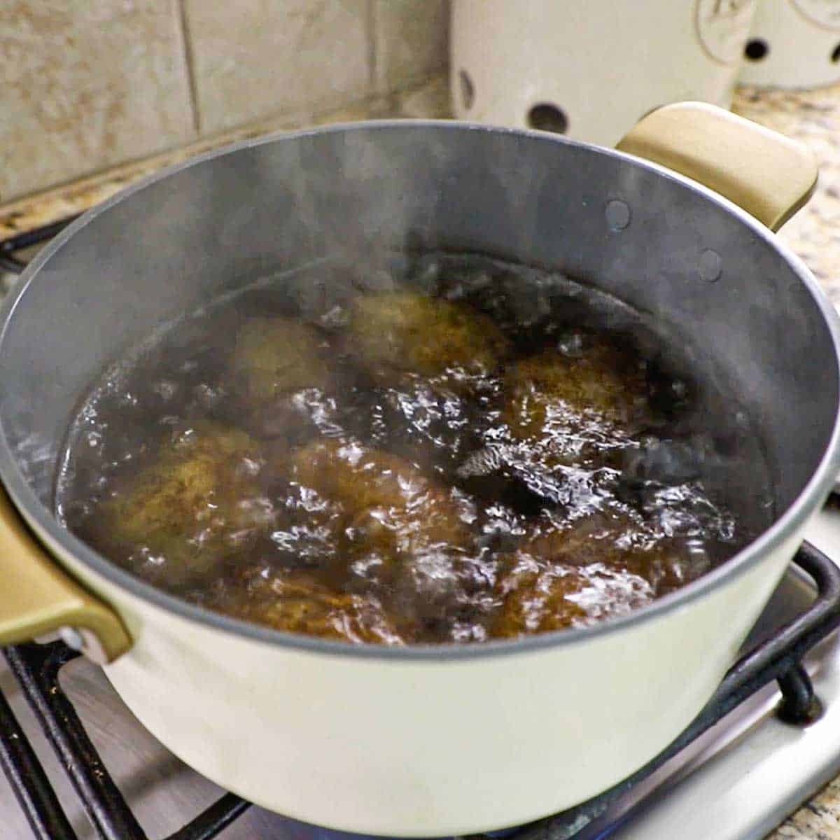 A large white pot on a gas stove filled with whole russet potatoes simmering in boiling water.