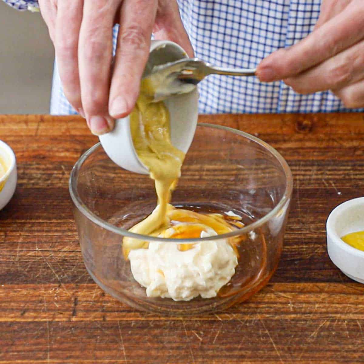 A person using a spoon to transfer Dijon mustard from a small bowl into a larger glass bowl containing mustard and honey.