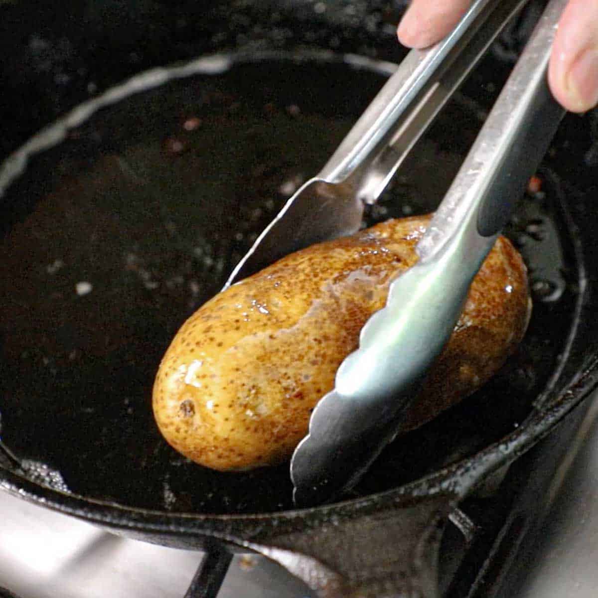 Person using a pair of metal tongs to drag an uncooked russet potato through rendered bacon grease in a case-iron skillet.