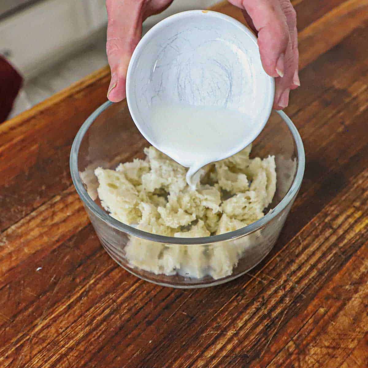 A person pouring whole milk from a small white bowl into a glass bowl filled with pieces of torn white bread.
