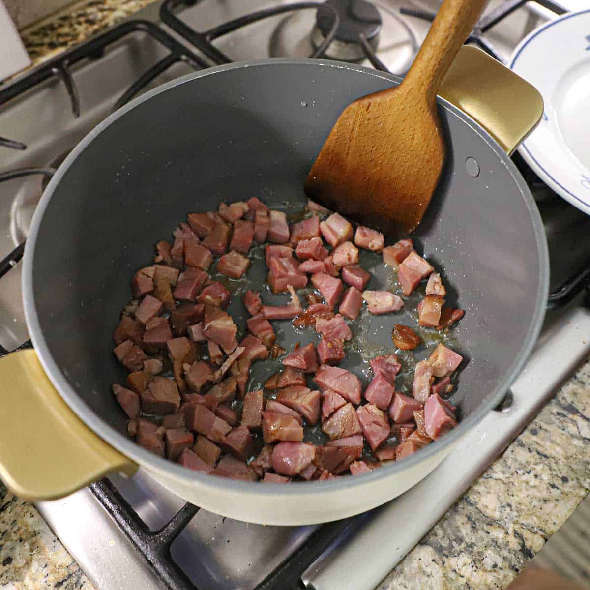 A person using a large wooden spatula to stir chunks of crispy ham that are being cooked in a large pot with hot oil.