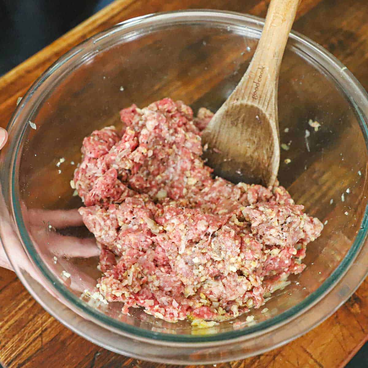 A person using a large wooden spoon to mix ground beef, ground pork, and other ingredients in a glass bowl for homemade Swedish meatballs.