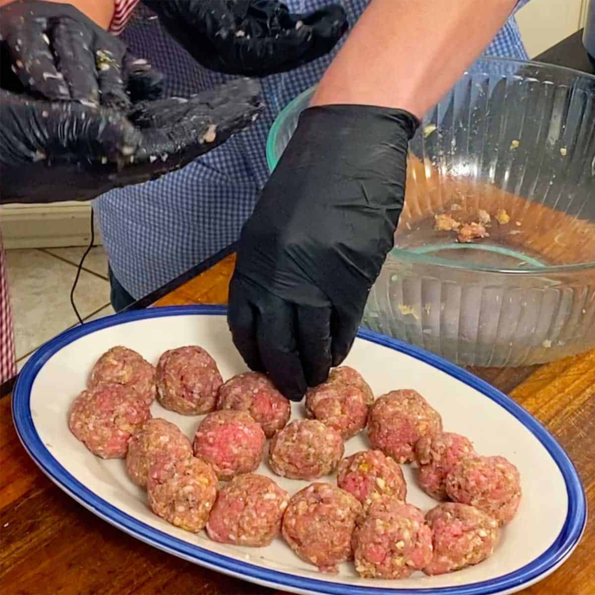 A person wearing black kitchen gloves forming uncooked Swedish meatballs and placing them on an oval platter.