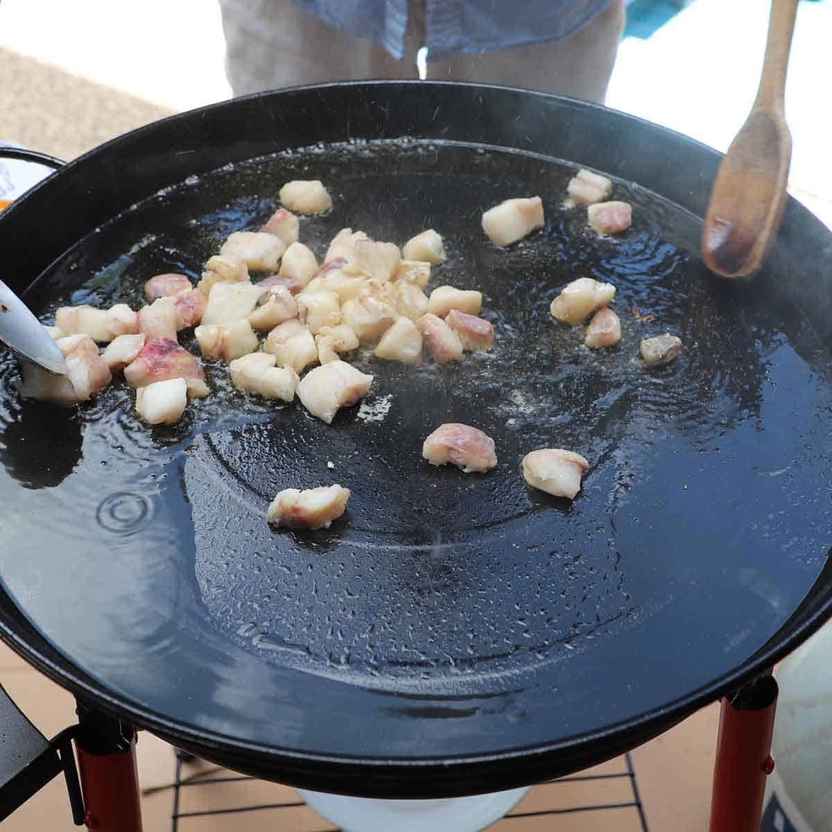 A person using a wooden spoon to toss chunks of monkfish in hot oil in a large paella pan.