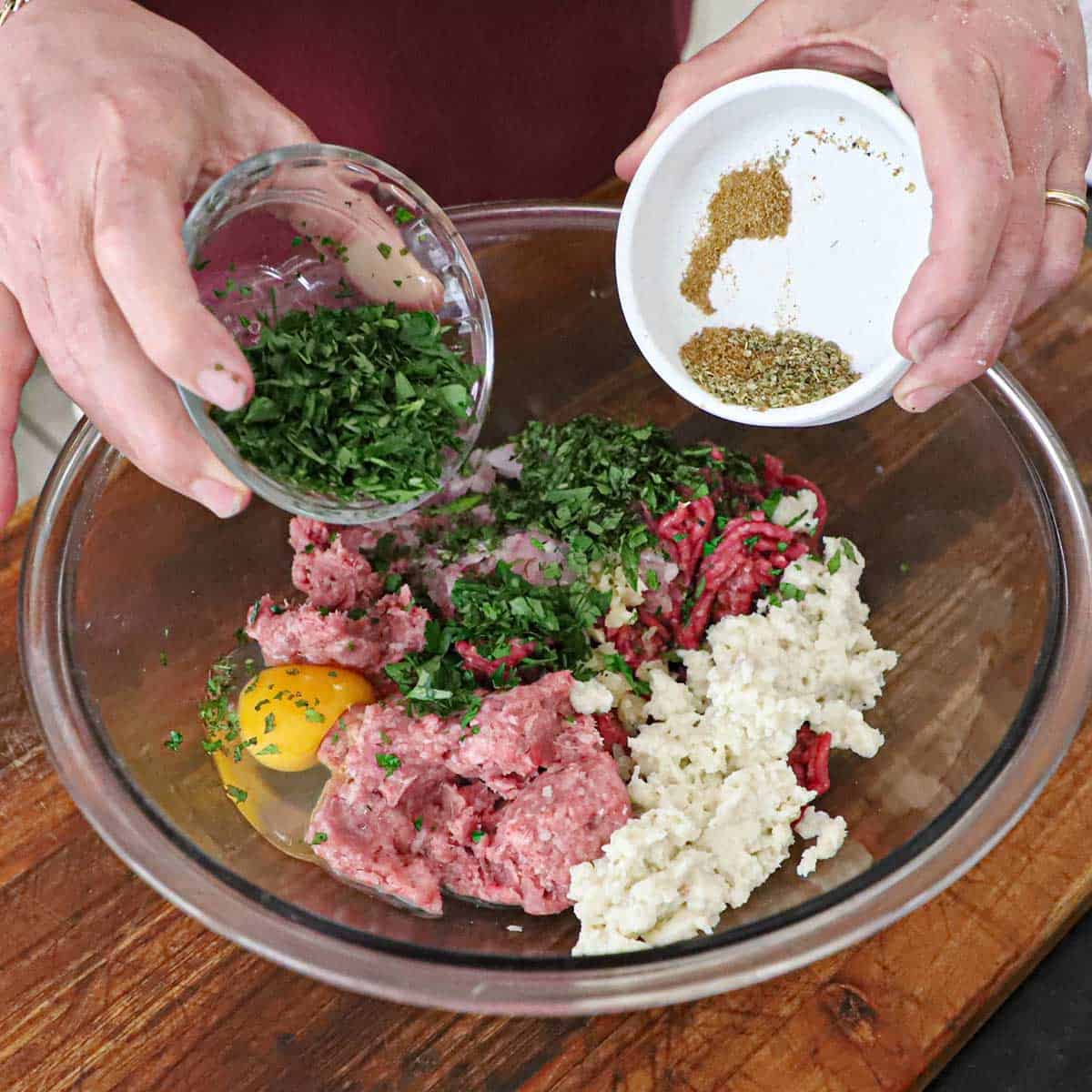A person holding a small glass bowl filled with chopped herbs in one hand and the other hand holding a small bowl of spices both over a glass bowl filled with the ingredients for Greek meatballs.