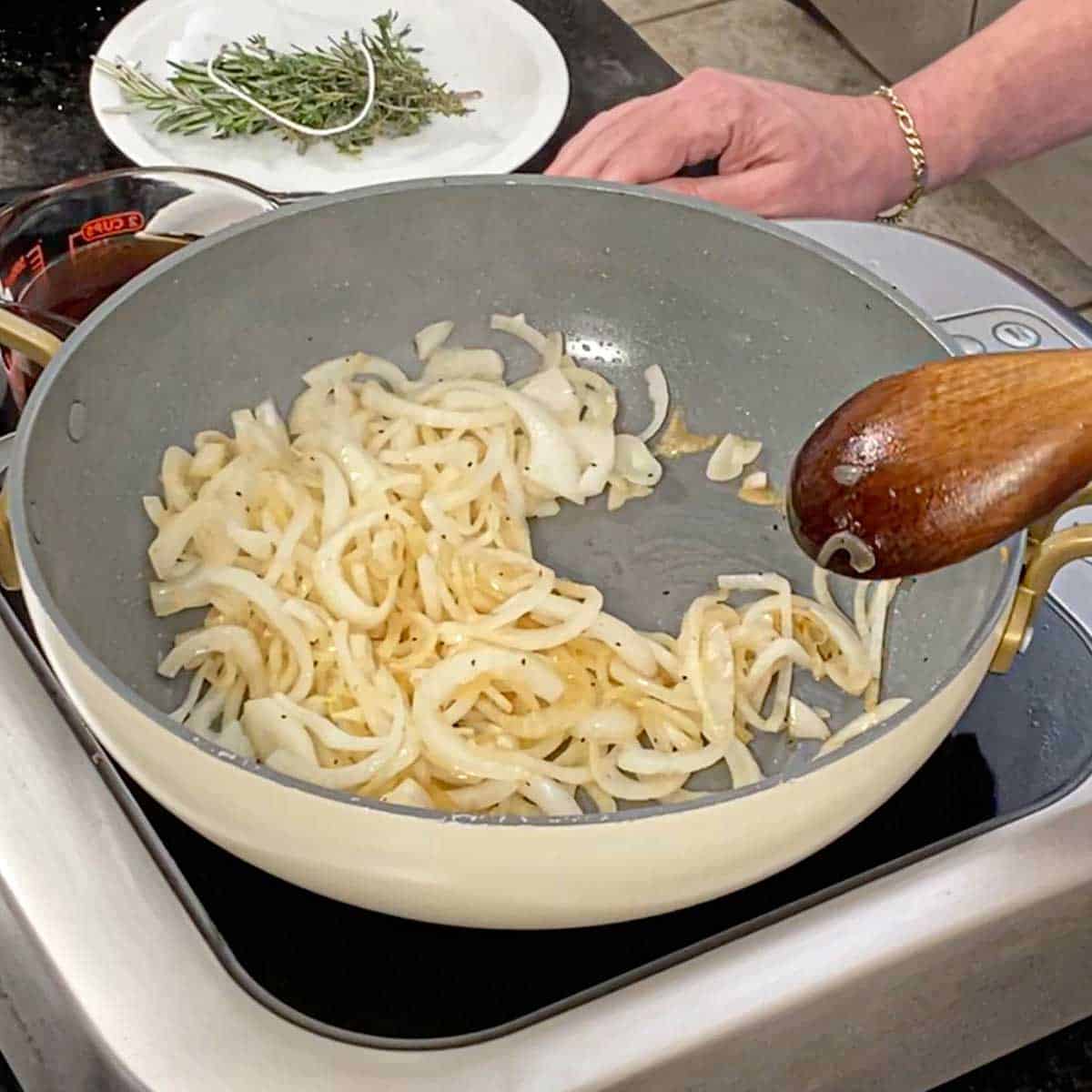 A person using a large wooden spoon to stir strips of onion that are being sautéed in a large non-stick skillet on an induction stovetop.