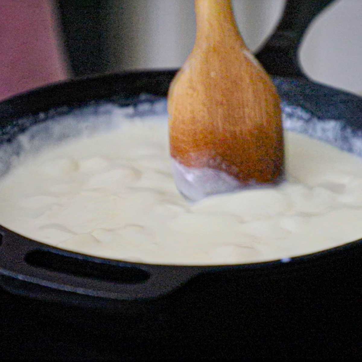 A person using a wooden spoon to stir melting white American cheese with milk and water in a black cast-iron skillet over low heat.