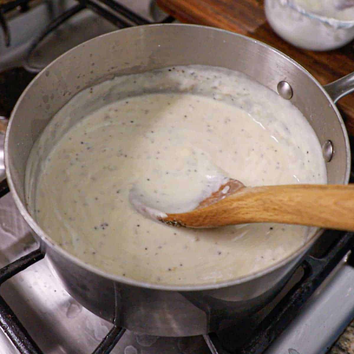 A person using a wooden spoon to stir a thickened cream of chicken and sour cream sauce in a saucepan on a gas stove.