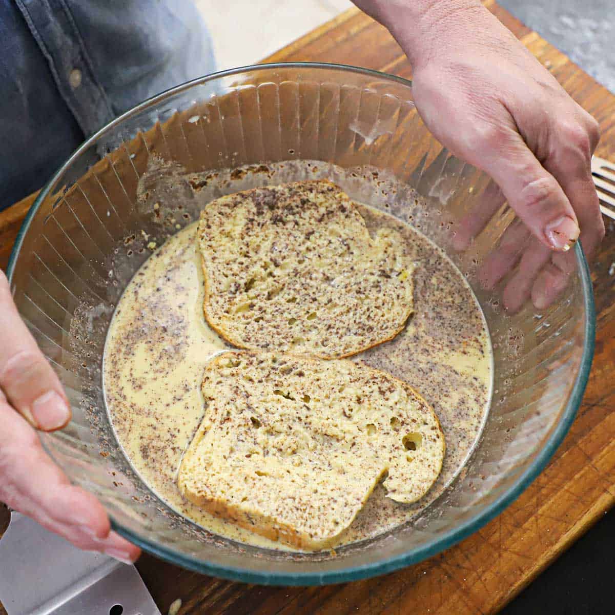 A person holding a large glass bowl that is filled with a French toast custard with two thick slices of brioche bread submerged in the liquid.